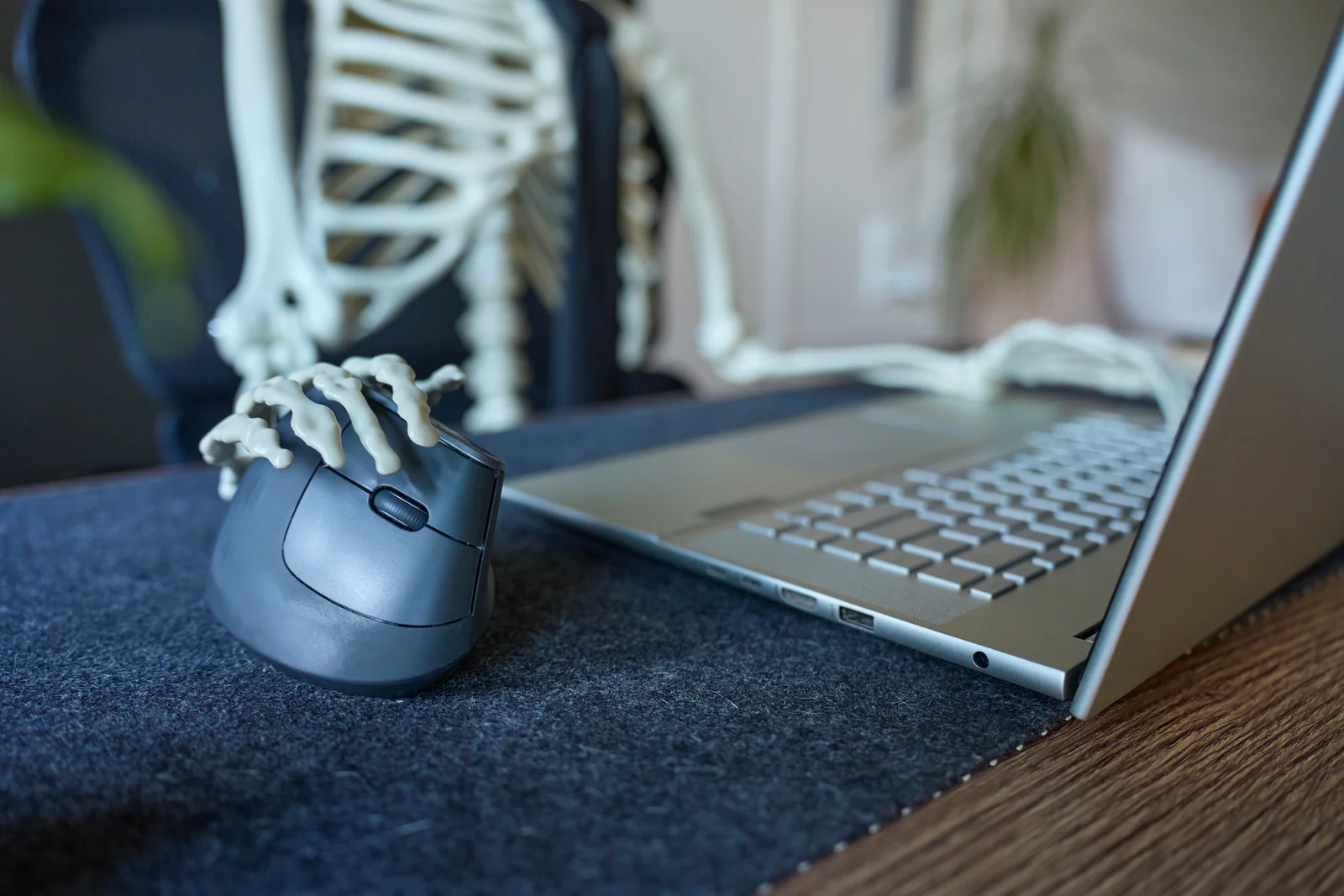 A skeleton seated at a desk with one bony hand on a computer mouse beside an open laptop